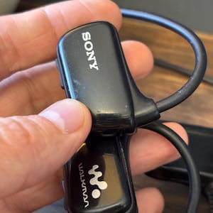 May include: A black Sony Walkman wearable digital music player featuring a curved, wrap-around design with the Sony logo printed in white on the top section and the Walkman logo printed in white on the lower section, including a black cable.