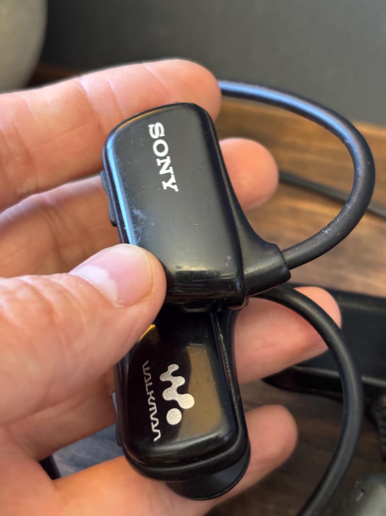 May include: A black Sony Walkman wearable digital music player featuring a curved, wrap-around design with the Sony logo printed in white on the top section and the Walkman logo printed in white on the lower section, including a black cable.