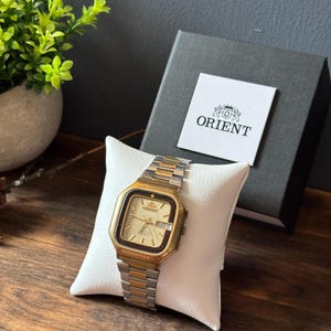 May include: A gold and silver-toned Orient wristwatch with a square face, a light-coloured dial, and a day-date display. The watch is displayed on a white cushion in front of a black Orient branded box with the Orient logo and text printed on it.
