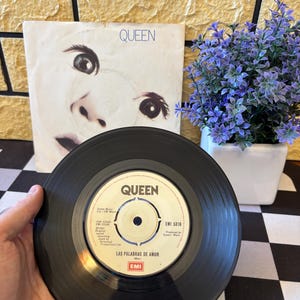 以下が含まれることがあります： 「LAS PALABRAS DE AMOR」の曲名がラベルに印刷された、Queenの黒いビニールレコード。背景には、顔とバンド名が描かれたアルバムカバーがあります。