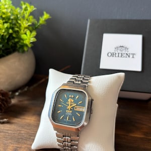 May include: A silver-toned Orient wristwatch with a square case and a dark blue dial. The watch features gold-colored hour markers, hands, and a day-date window at the three o'clock position. It has a metal link bracelet and is displayed on a white pillow.