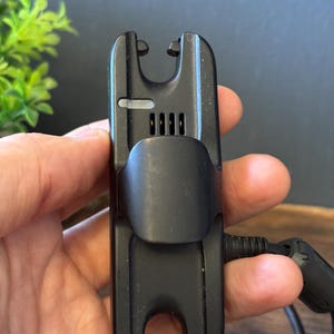 May include: A black plastic electronic device featuring a central clip, a small rectangular light indicator, and four vertical ventilation slots. The device has a U-shaped cutout at both the top and bottom ends and includes a black power cable.