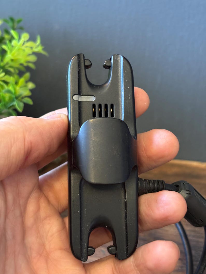 May include: A black plastic electronic device featuring a central clip, a small rectangular light indicator, and four vertical ventilation slots. The device has a U-shaped cutout at both the top and bottom ends and includes a black power cable.