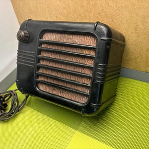 May include: A vintage black radio with a brown speaker grille and a knob on the side. The radio is plugged in with a black cord.