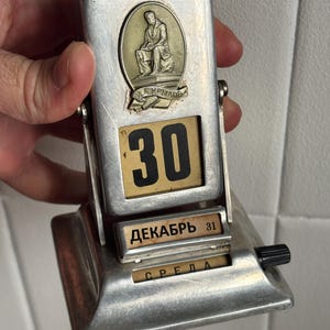 May include: A silver-toned metal perpetual desk calendar featuring a central medallion of a seated figure with the text "И.А.КРЫЛОВ". The display shows the number 30, the month "ДЕКАБРЬ 31", and the day "СРЕДА" in Cyrillic script with a black adjustment knob.