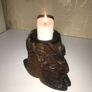 May include: A wooden candle holder carved in the shape of a face with intricate details. A white candle is placed in the holder and is lit.