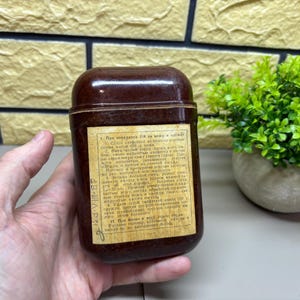 Op de afbeelding: Een vintage, rechthoekige, donkerbruine container met een afgeronde bovenkant. Een label met tekst is op de voorkant bevestigd. De container wordt in een hand gehouden, met een kleine groene plant op de achtergrond.