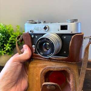 May include: A vintage FED 3 camera with a silver body and black accents, housed in a brown leather case. The lens is prominently displayed, and the camera has a classic, retro aesthetic. The case has a strap.