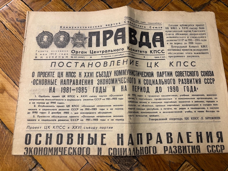May include: A black and white newspaper headline reads "POSTANOVLENIE CK KPSS" and "OSNOVNYE NAPRAVLENIYA EKONOMICHESKOGO I SOCIAL'NOGO RAZVITIYA SSSR NA 1981-1985 GODY I NA PERIOD DO 1990 GODA". The newspaper is titled "PRAVDA" and is the "Organ Tsentral'nogo Komiteta KPSS".