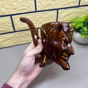 Puede incluir: Una taza de cerámica marrón con forma de diablo con un asa de cola curvada. La taza presenta una cara detallada con cuernos y una expresión sonriente. La taza está esmaltada con un acabado brillante.