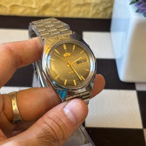 Puede incluir: Un reloj Orient plateado con esfera dorada y visualización de fecha "MON 28". El reloj tiene una correa plateada y lo sostiene una mano. La esfera del reloj tiene agujas y marcadores dorados.