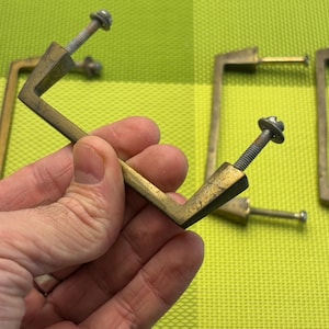 Pode incluir: Quatro puxadores de porta de armário de latão com forma quadrada e extremidade pontiaguda. Os puxadores têm um acabamento escovado e são presos por parafusos.