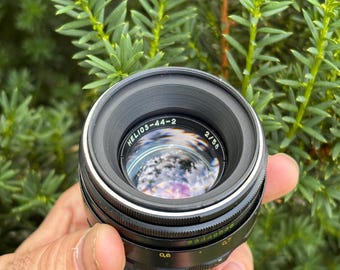 Może przedstawiać: Czarny obiektyw fotograficzny Helios-44-2 2/58 z okrągłym elementem szklanym. Na tubusie obiektywu znajduje się biały napis HELIOS-44-2 2/58, zielone oznaczenia numeryczne oraz czarny dekielek u podstawy, z widocznymi odbiciami w szkle.