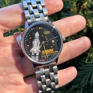 May include: A vintage wristwatch with a silver-tone metal band and a black face. The watch face features a white and yellow illustration of a spacecraft, with the Cyrillic word "Buran" in yellow. The watch is held in a hand.