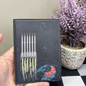 May include: Black book with a space-themed design. The cover features rockets ascending into space above a globe. The globe is blue and red, with stars in the background. The book is held in a hand.