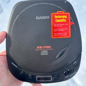 May include: A black Casio PZ-700 portable compact disc player with a bass boost system. It has a digital display and control buttons for program, mode, stop, and play/pause. A red sticker indicates recharging capability for NiCd batteries.