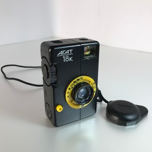 以下が含まれることがあります： 黄色いダイヤルとレンズリングが付いた黒いAgat 18Kフィルムカメラ。カメラには黒いストラップと黒いレンズキャップが付いています。「AGAT 18K」の文字がカメラの前面に印刷されています。
