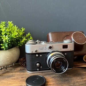 May include: A silver and black FED 3 rangefinder camera with a lens marked 2.8/52 and I-61. The camera features a textured black grip, a top-mounted film advance lever, and a shutter speed dial. A separate black circular lens cap sits in front.