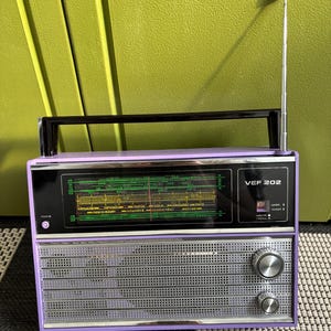 Puede incluir: Una radio vintage VEF 202 de color morado con una rejilla de altavoz plateada y una perilla negra. La radio tiene una pantalla digital que muestra "KBI".