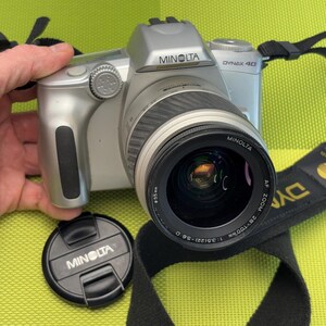 以下が含まれることがあります： 黒いレンズと黒いストラップが付いた銀色のミノルタダイナックス40カメラ。カメラにはミノルタのロゴが付いたレンズキャップが付いています。