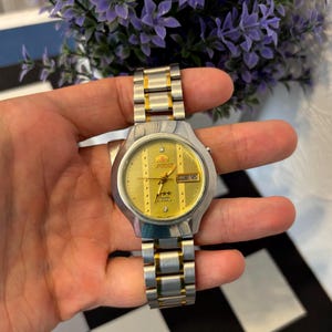 May include: A silver and gold-toned Orient watch with a gold dial. The watch features a date display and a silver and gold-toned band. The watch is held in a hand.