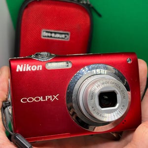以下が含まれることがあります： 前面に「COOLPIX」と「Nikon」の文字がプリントされた、赤いニコンクールピクスデジタルカメラ。シルバーのレンズが付いています。カメラにはリストストラップと赤いカメラケースが付いています。