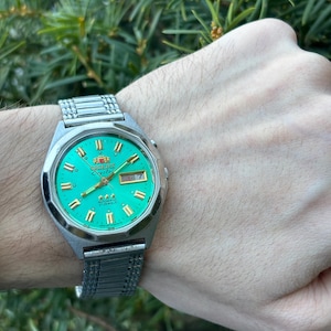 May include: A vintage Orient Crystal watch with a vibrant green face and gold-tone accents. The watch features a silver-tone stainless steel band and a day-date display. The watch is 37mm in diameter.