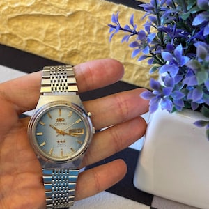 May include: A silver-toned Orient wristwatch with a light blue face and a metal band. The watch face displays the day and date, along with the Orient logo and three stars. The watch is held in a hand.