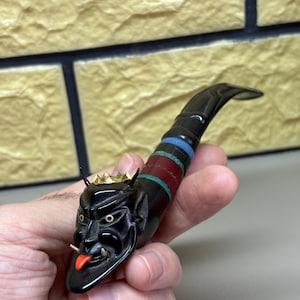 May include: A black tobacco pipe with a devil face design. The pipe has a red tongue, gold crown, and a striped stem with blue, red, and green bands. The pipe is held in a hand.