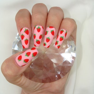 May include: Pink long, almond-shaped fake nails with a strawberry print design.