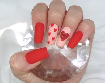 May include: A hand displaying a set of red and pink press-on nails. The nails are square-shaped with a matte finish. Some nails are solid red, while others feature small red hearts or a glittery heart design.