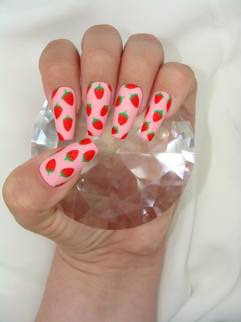 May include: Pink long, almond-shaped fake nails with a strawberry print design.