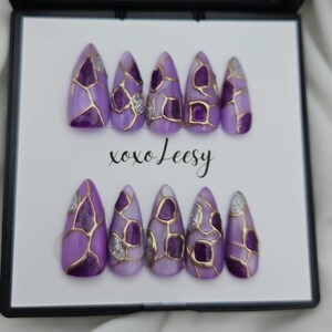 May include: Set of ten lavender-colored press-on nails with a gold geometric design and glitter accents. The nails are almond-shaped and feature the text "xoxo Leesy".