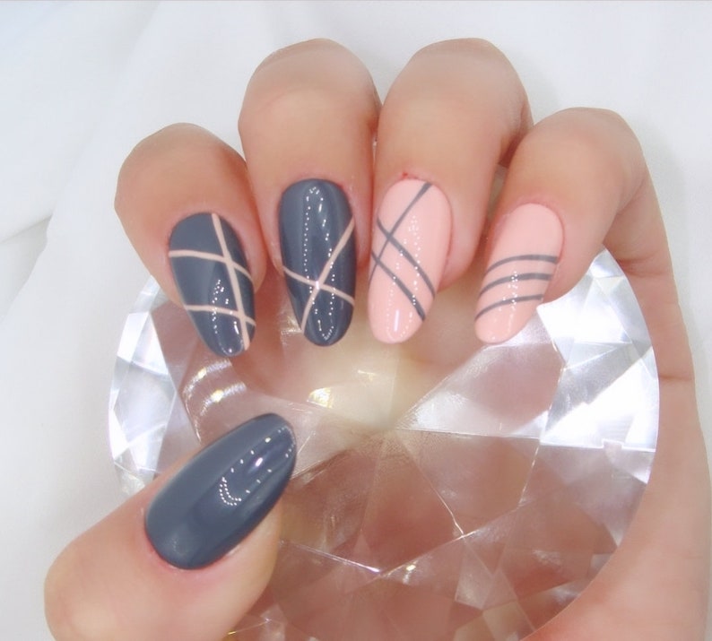 May include: Five oval-shaped nails with a criss-cross design. Two nails are painted a light pink color with dark gray criss-cross lines. Three nails are painted a dark gray color with light pink criss-cross lines.