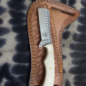 May include: A small, silver knife with a white handle and gold accents, resting in a brown leather sheath. The sheath has decorative stitching and a textured pattern. The knife blade has a serrated edge.