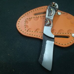 May include: A small, handcrafted knife with a marbled white, brown, and black handle. The blade is silver and has a curved edge. It rests on a brown leather sheath with decorative stitching and a snap closure. The knife is designed for utility.