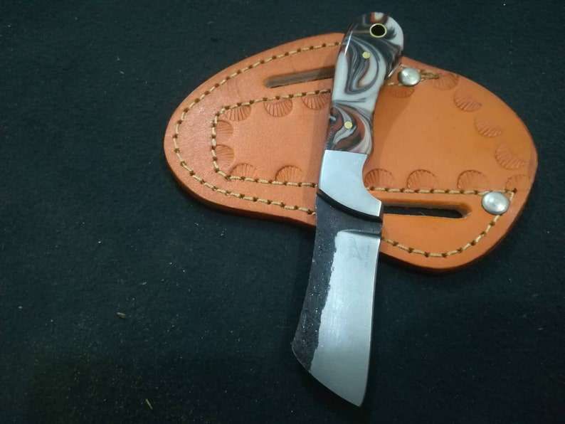 May include: A small, handcrafted knife with a marbled white, brown, and black handle. The blade is silver and has a curved edge. It rests on a brown leather sheath with decorative stitching and a snap closure. The knife is designed for utility.