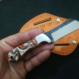 May include: A small, handmade knife with a marbled orange, black, and white handle. The blade is silver and the knife rests on an orange leather sheath with decorative stitching. The knife is held in a hand.