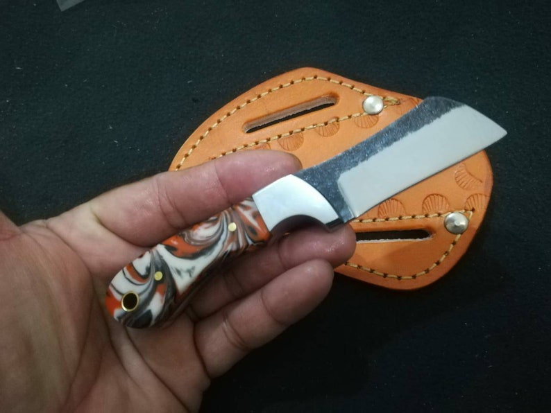 May include: A small, handmade knife with a marbled orange, black, and white handle. The blade is silver and the knife rests on an orange leather sheath with decorative stitching. The knife is held in a hand.