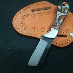 May include: A small, handcrafted knife with a polished, curved blade and a handle featuring red, white, and black swirls. The knife rests on a tan leather sheath with decorative stitching and a snap closure. The blade has a dark, textured finish.