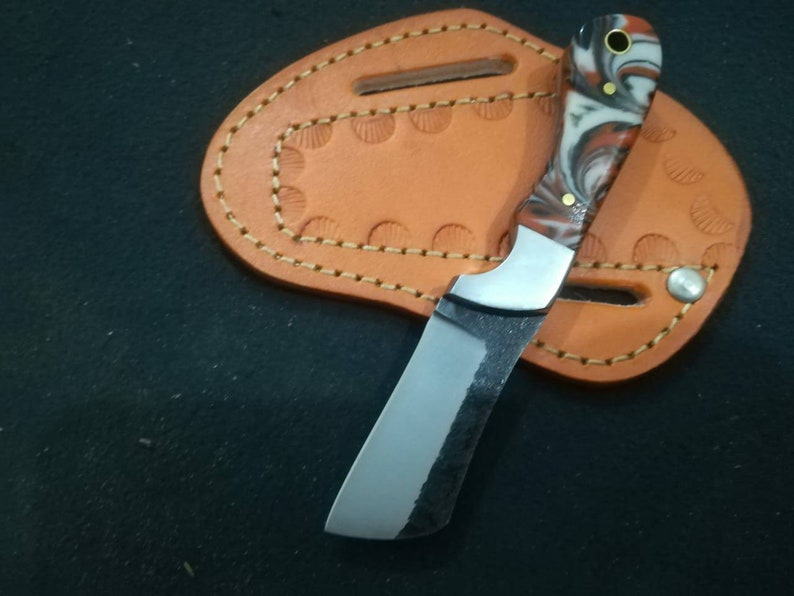 May include: A small, handcrafted knife with a polished, curved blade and a handle featuring red, white, and black swirls. The knife rests on a tan leather sheath with decorative stitching and a snap closure. The blade has a dark, textured finish.