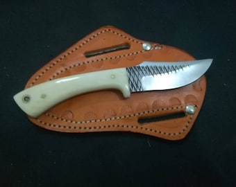 May include: A handcrafted knife with a cream-colored handle and a silver blade, resting on a brown leather sheath. The blade has a decorative etched pattern near the handle. The sheath has a decorative stitched border.
