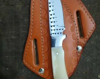 May include: A handcrafted knife with a stainless steel blade and a cream-colored handle. The blade has a decorative triangular pattern. The knife rests in a tan leather sheath with decorative stitching and metal rivets.