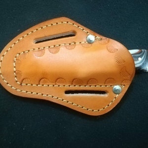 May include: A small, handcrafted knife with a marbled handle is partially inserted into a tan leather sheath. The sheath features decorative stitching and a scalloped edge design. Two silver rivets secure the sheath.
