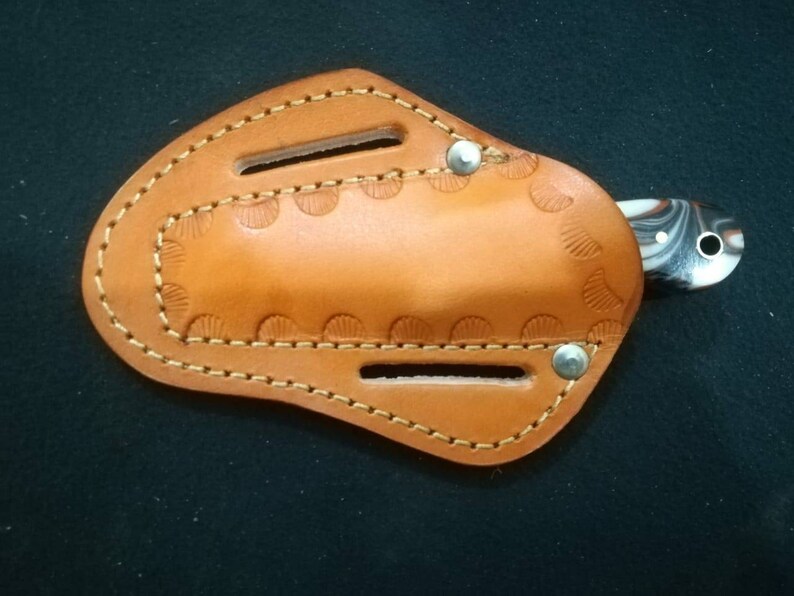 May include: A small, handcrafted knife with a marbled handle is partially inserted into a tan leather sheath. The sheath features decorative stitching and a scalloped edge design. Two silver rivets secure the sheath.
