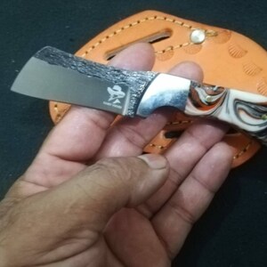 May include: A small, stainless steel knife with a textured blade and a handle featuring a swirl pattern of white, orange, and black. The knife has a silver bolster and a small logo. The knife is resting on a brown leather sheath.