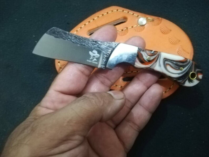 May include: A small, stainless steel knife with a textured blade and a handle featuring a swirl pattern of white, orange, and black. The knife has a silver bolster and a small logo. The knife is resting on a brown leather sheath.