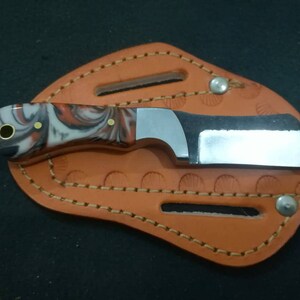 May include: A small, stainless steel knife with a marbled red, white, and black handle, resting in a brown leather sheath. The sheath has decorative stitching and is secured with metal rivets. The knife blade is approximately 2 inches long.