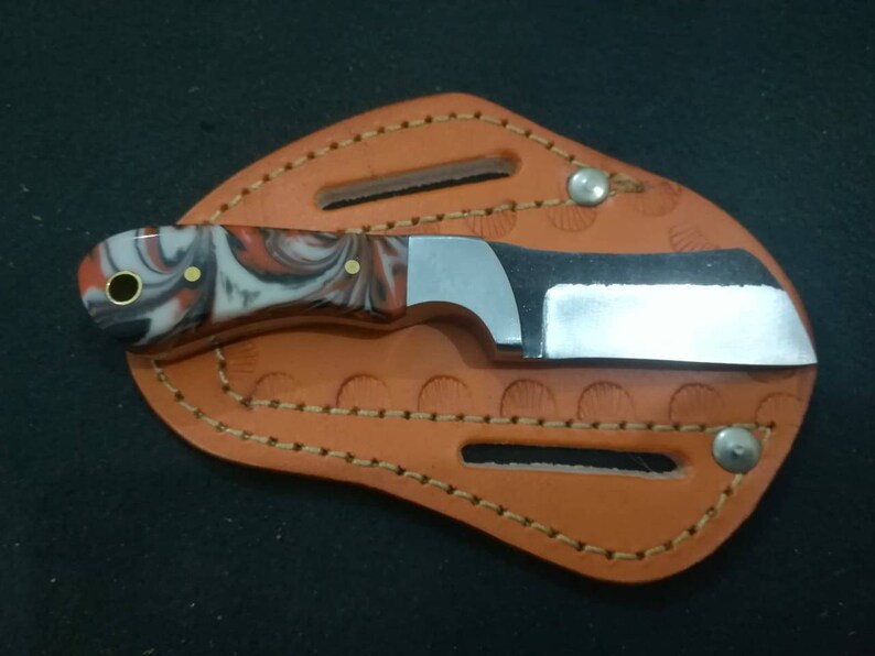 May include: A small, stainless steel knife with a marbled red, white, and black handle, resting in a brown leather sheath. The sheath has decorative stitching and is secured with metal rivets. The knife blade is approximately 2 inches long.