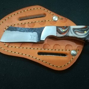 May include: A small, stainless steel knife with a marbled white, black, and brown handle, resting in a tan leather sheath. The blade has a dark, textured edge and the text "Eagle Knife".